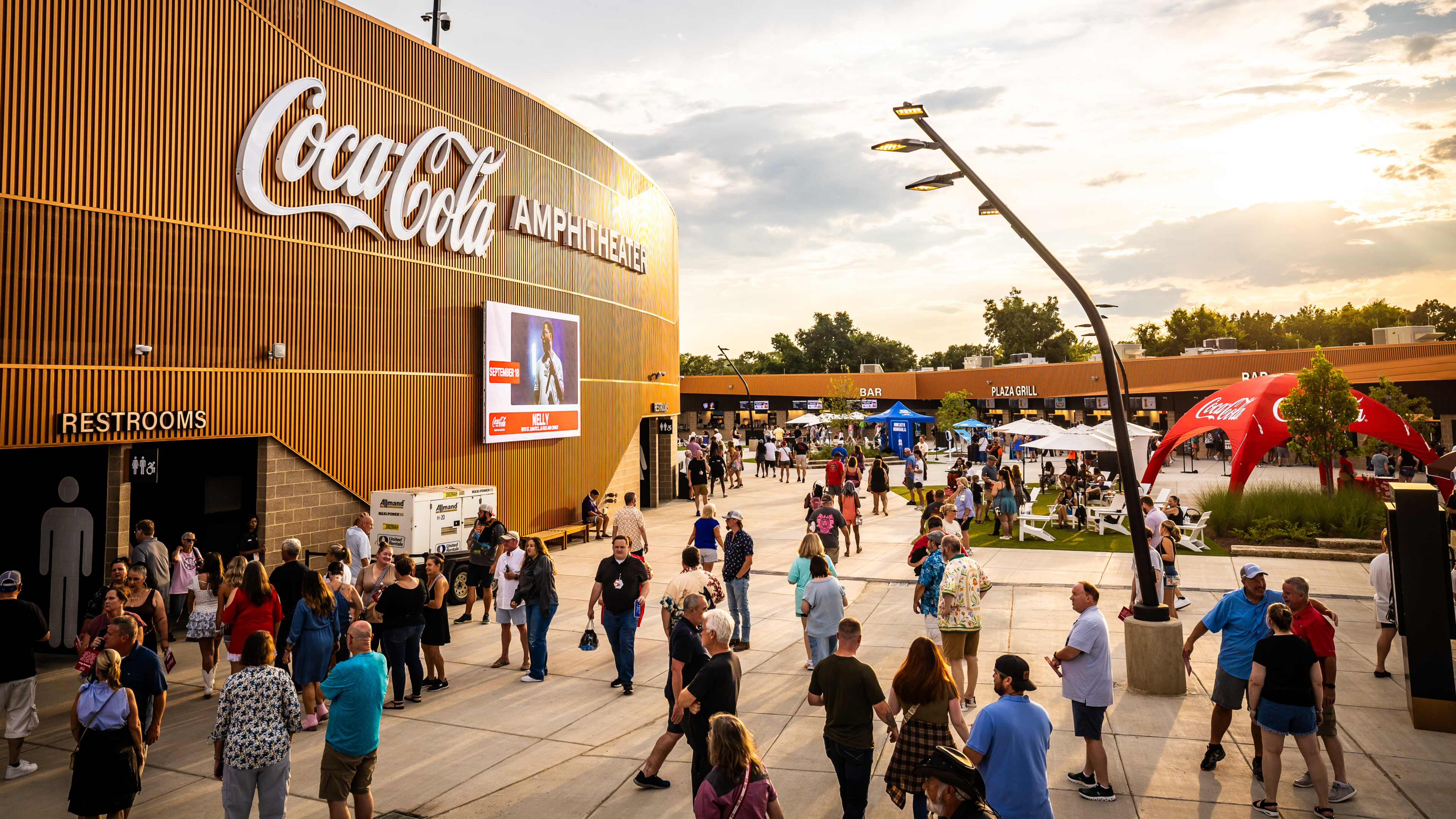 People gather in the plaza outside an outdoor amphitheater at sunset, with venue signage, food and drink areas, and landscaped seating.