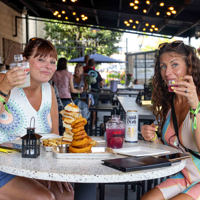 Two woman holding up their drinks (shaker cup cocktails) for the camera with chicken sliders and onion rings on their table, in our outdoor VIP club area