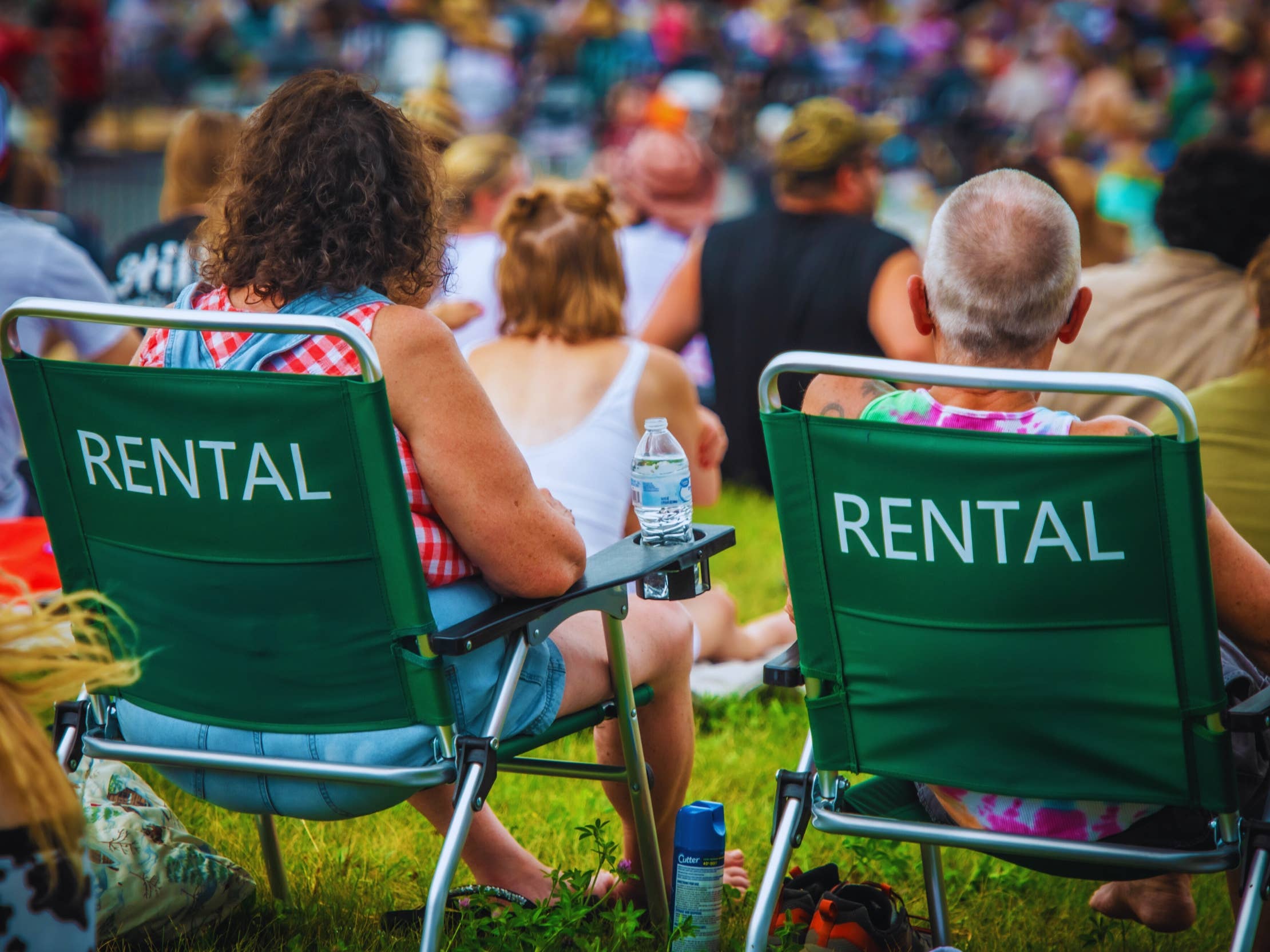 Two people are seen from behind while sitting in rental lawn chairs on the grass.