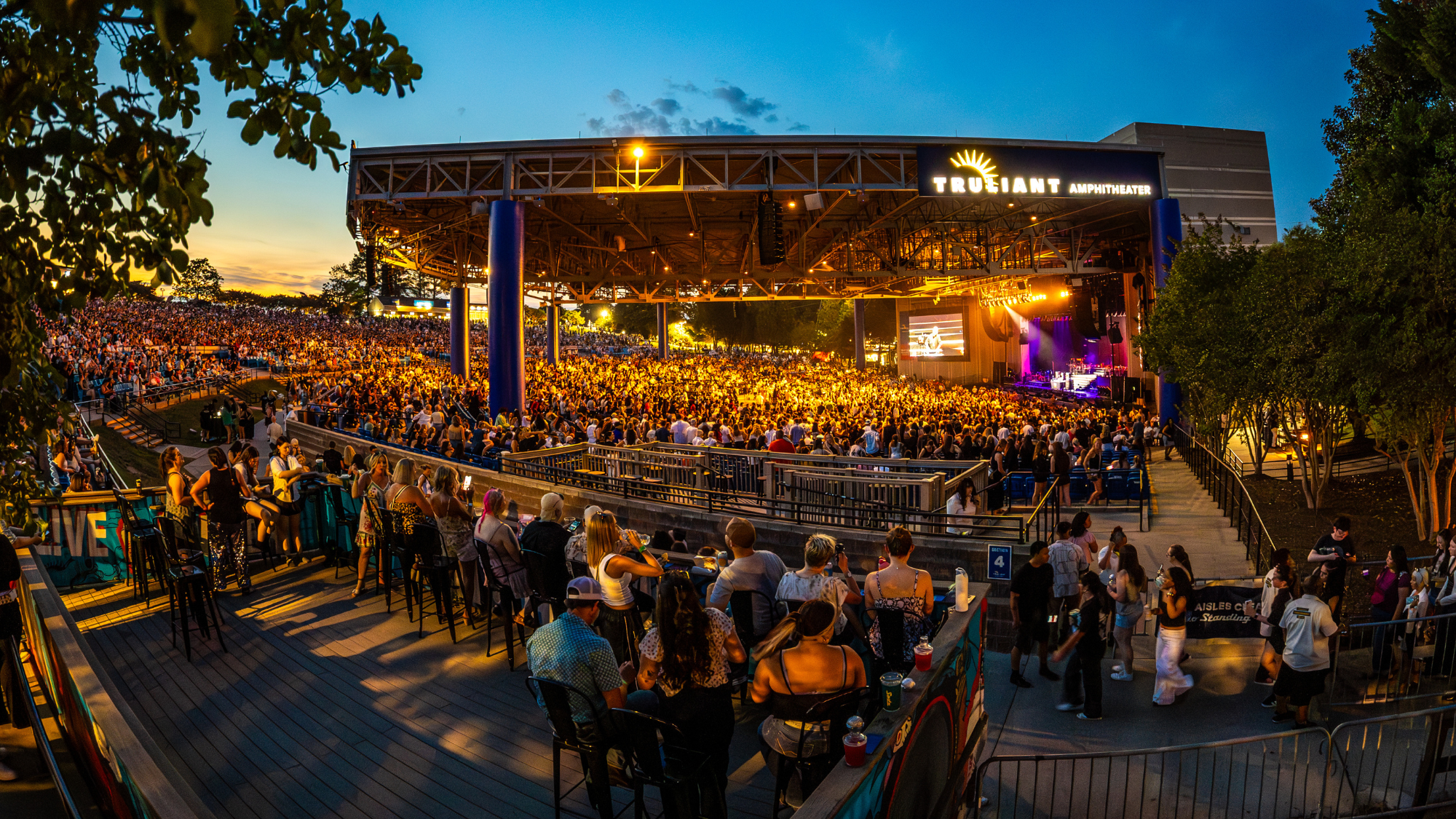A wide shot of the viewing deck in section 4. Fans are sitting in chairs on the deck with a side view of the concert. Stage is glowing, crowd is full, and sun is going down.