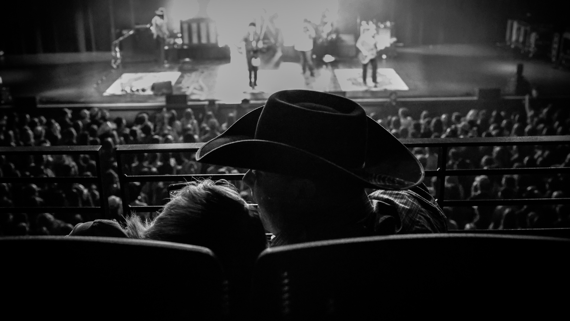A man and a woman sitting in the audience at a concert with her head on his shoulder.