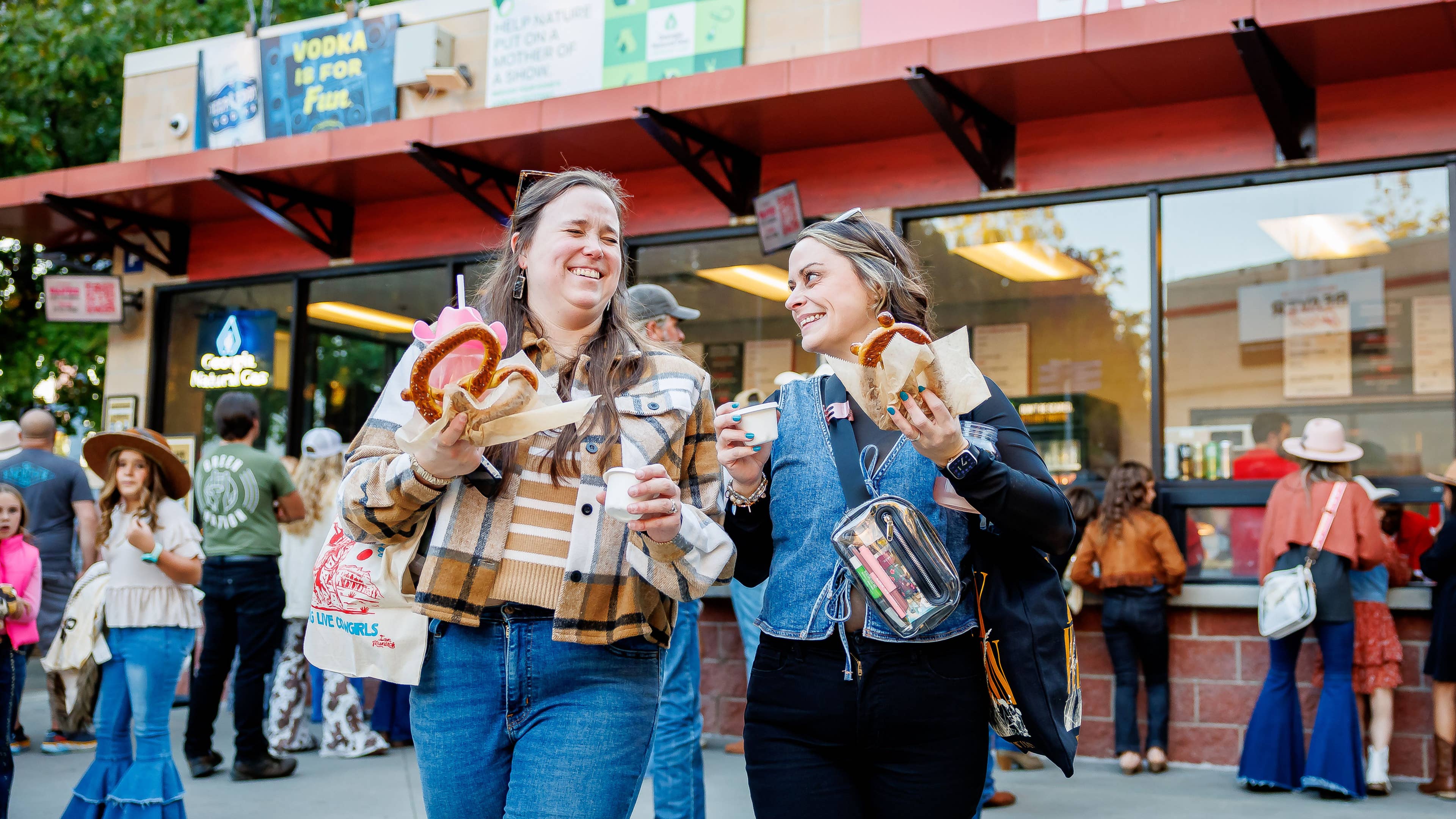 Two friends smiling and holding pretzels in front of the concession stands