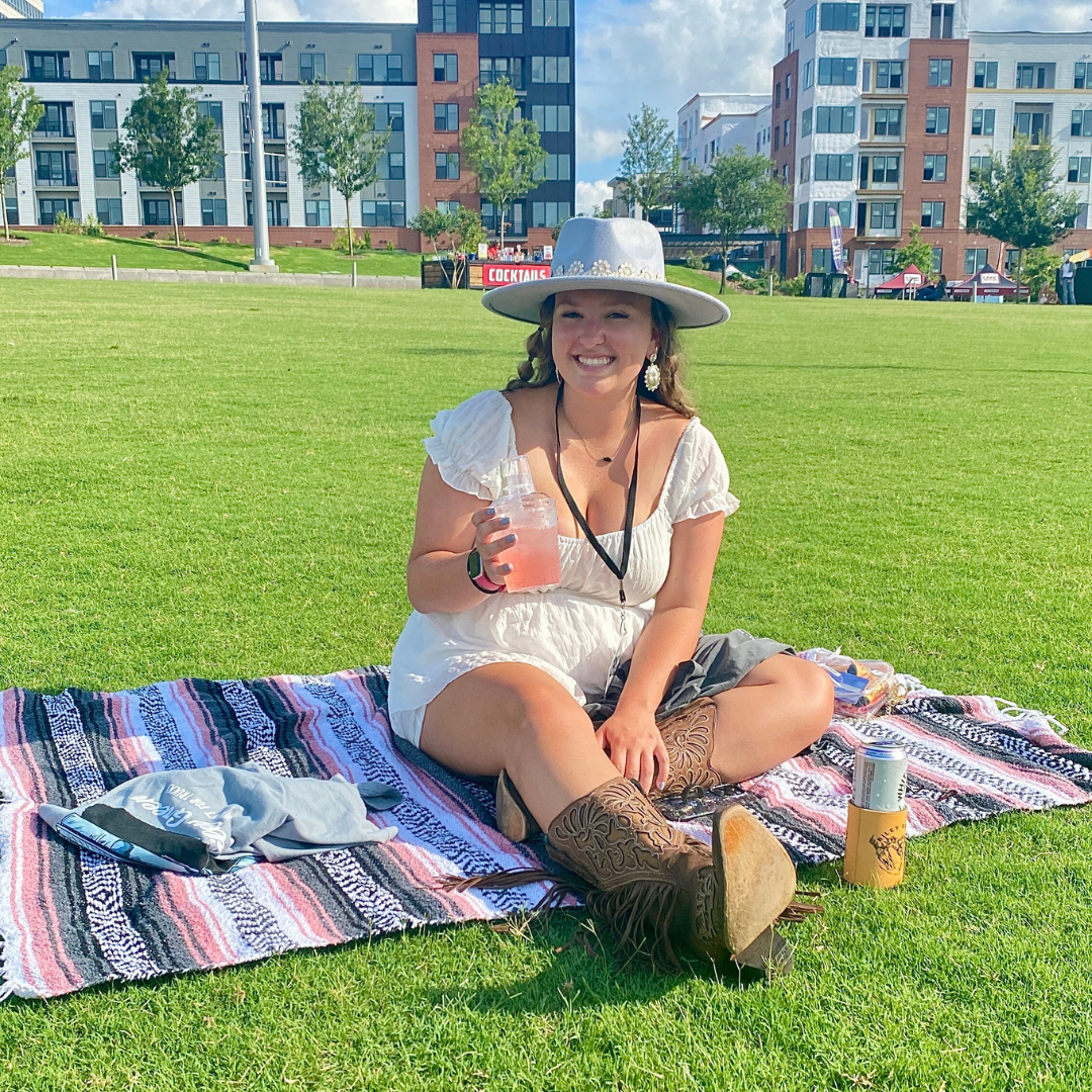 A girl sitting on a blanket in the lawn holding a drink. 