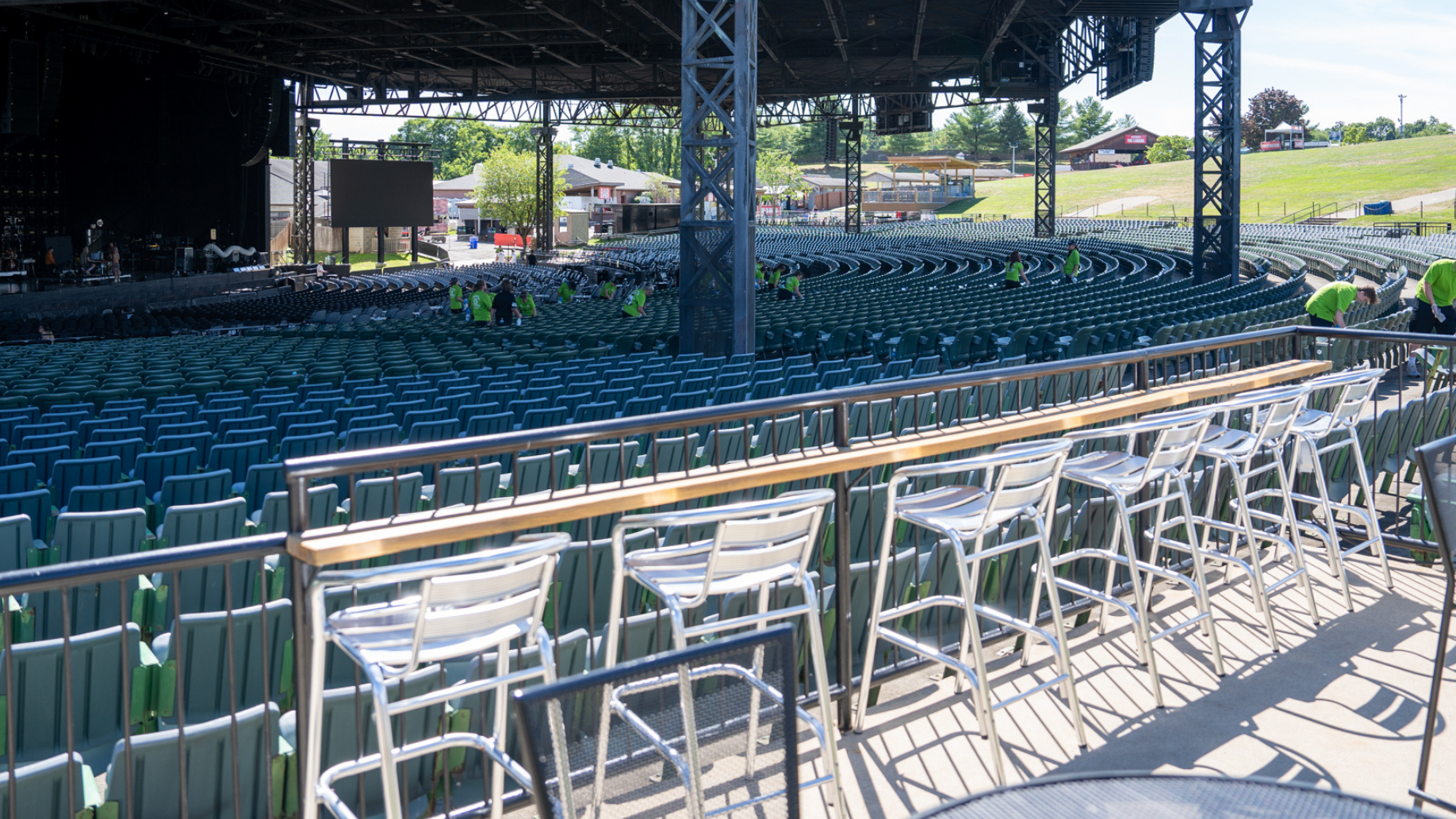 A row of empty high top chairs along a railing with a high-top table. Concourse section and stage in front of the table and chairs. 