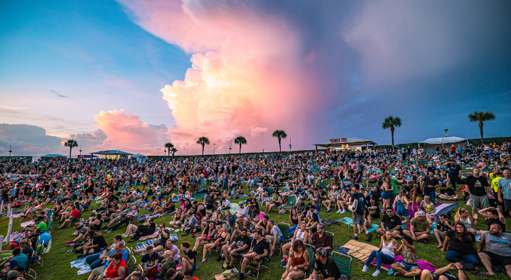 Large outdoor crowd on the lawn at MIDFLORIDA Credit Union Amphitheatre under a sunset sky.