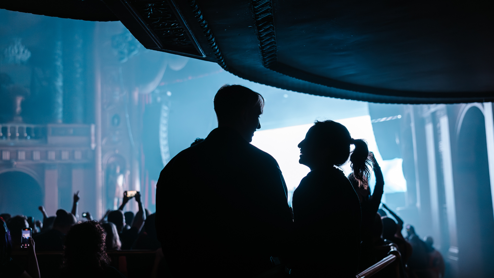 A couple looking at each other, smiling, as they stand in the ADA section of the venue. There is a concert happening behind them.