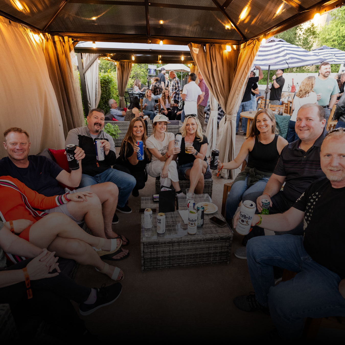 Fans sitting under a tented area with string lights smiling at the camera and holding their drinks up