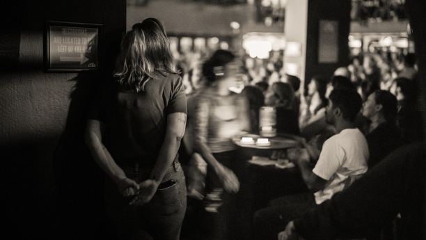 A woman looking towards the audience in the showroom, while a waitress walks by carrying drinks.