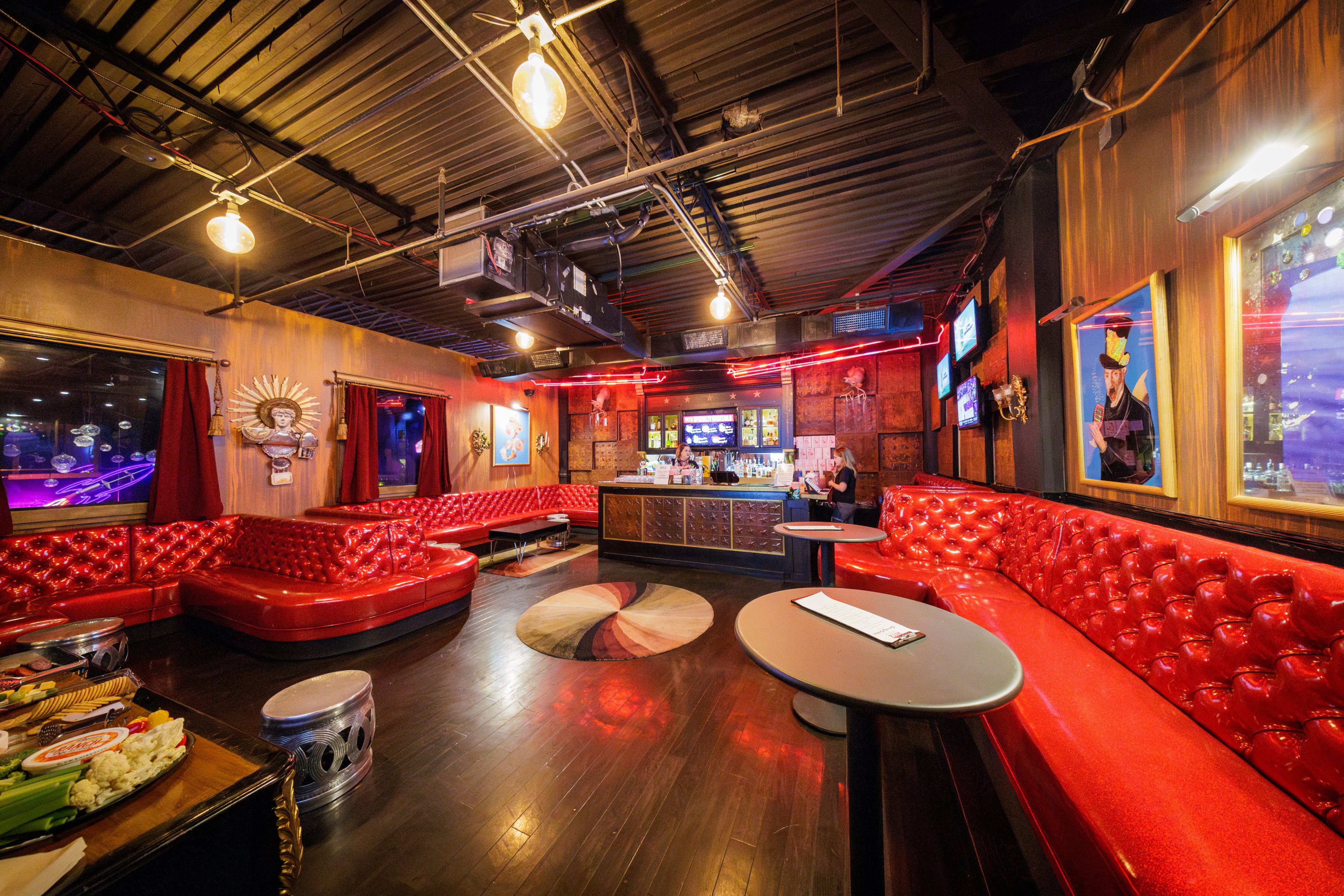 A small room with a bar with red shiny couches and tall bar tables