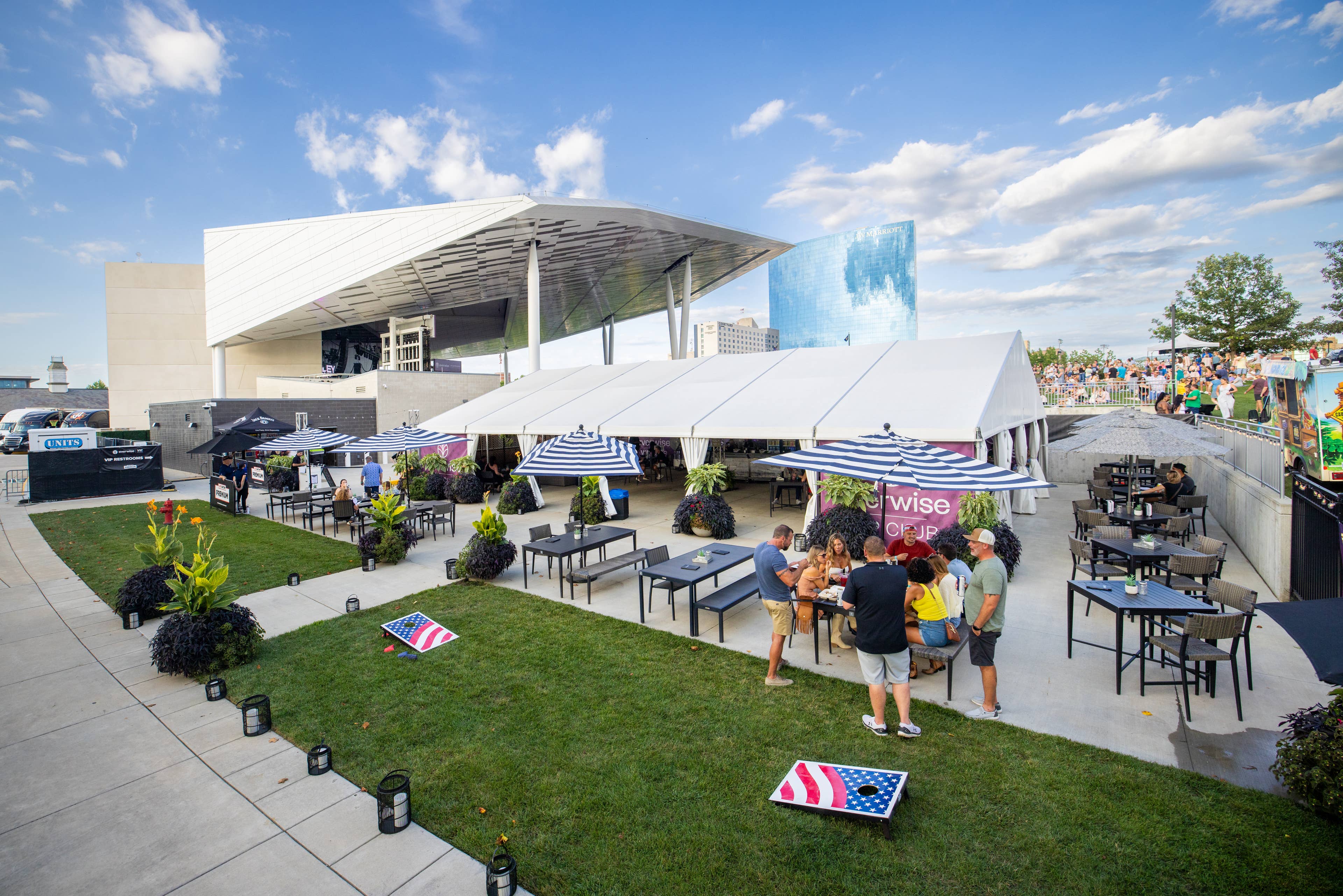 This photo shows the VIP club area. There is a group of people sitting at a table with an umbrella open over them. It shows the grassy areas, yard games and additional table and chairs.

Live Nation presents Jordan Davis at Everwise Amphitheater at White River State Park in Indianapolis, Indiana on July 31, 2024. (Photo by Alive Coverage for Live Nation)