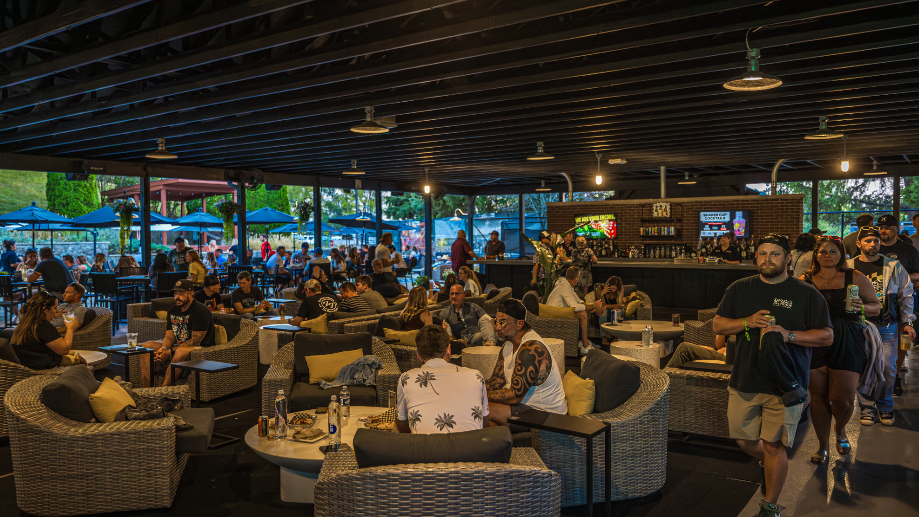 Multiple fans sitting, drinking, eating, and chatting in comfortable lounge chairs in a shaded area, with some fans standing near a private bar.
