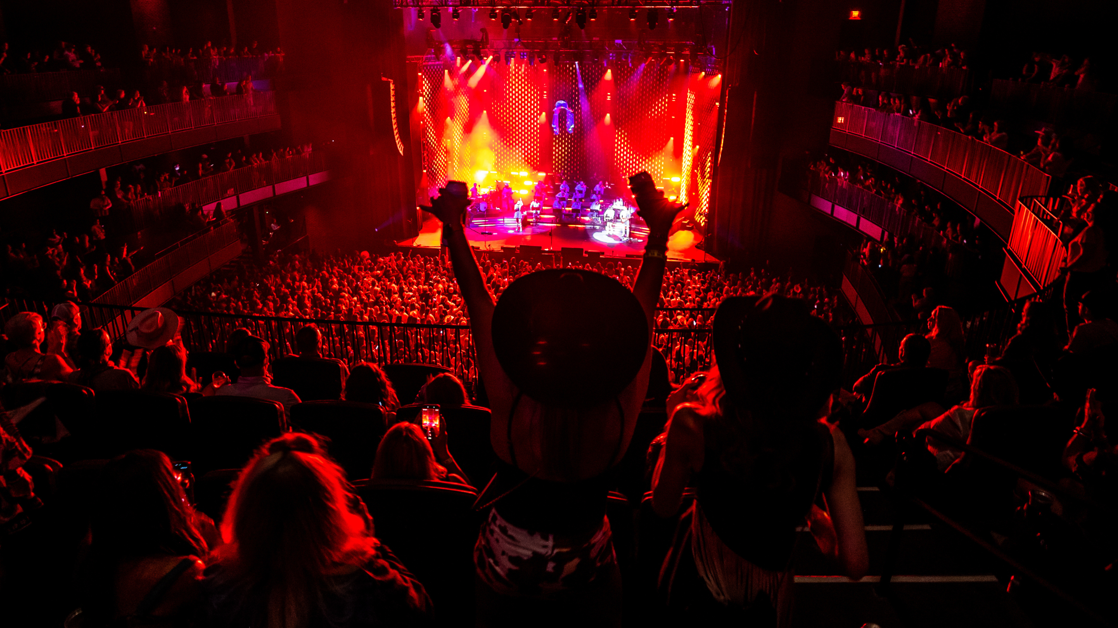 Concertgoers watch a live performance from an upper-level balcony as the stage glows red and gold. Photo credit: Alive Coverage