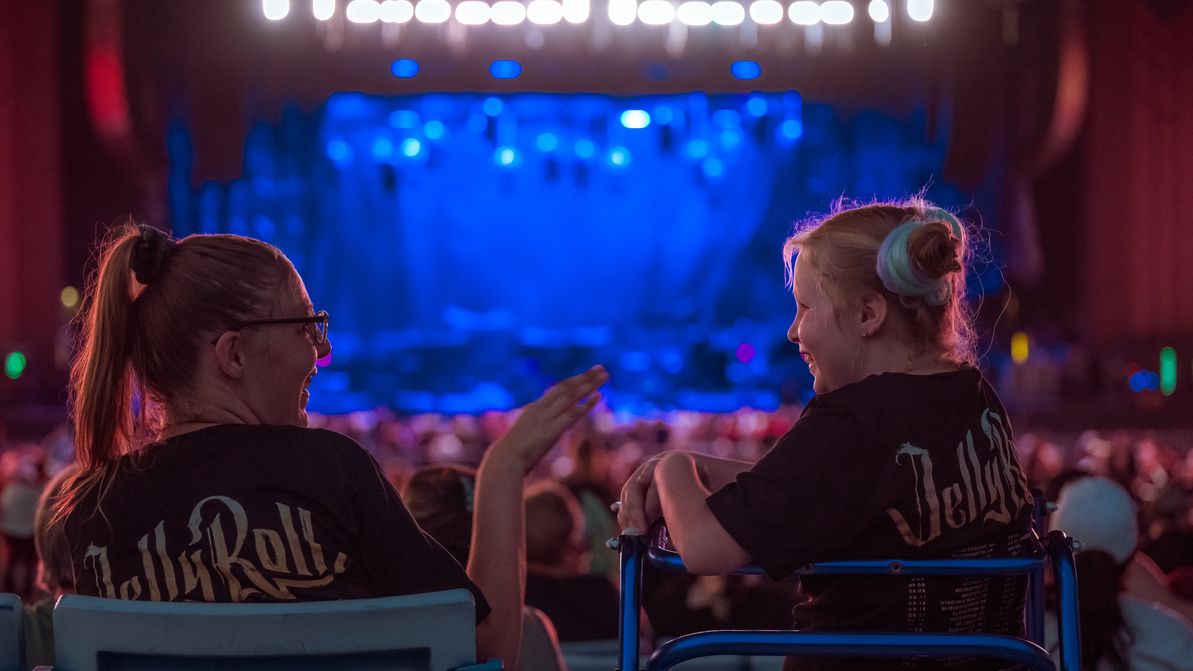 two fans smiling and talking to each other with the stage in the background