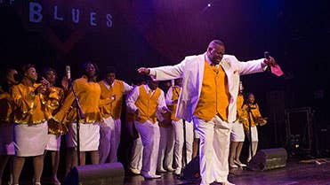 A group of performers in white suits and yellow shirts dancing and singing on stage

