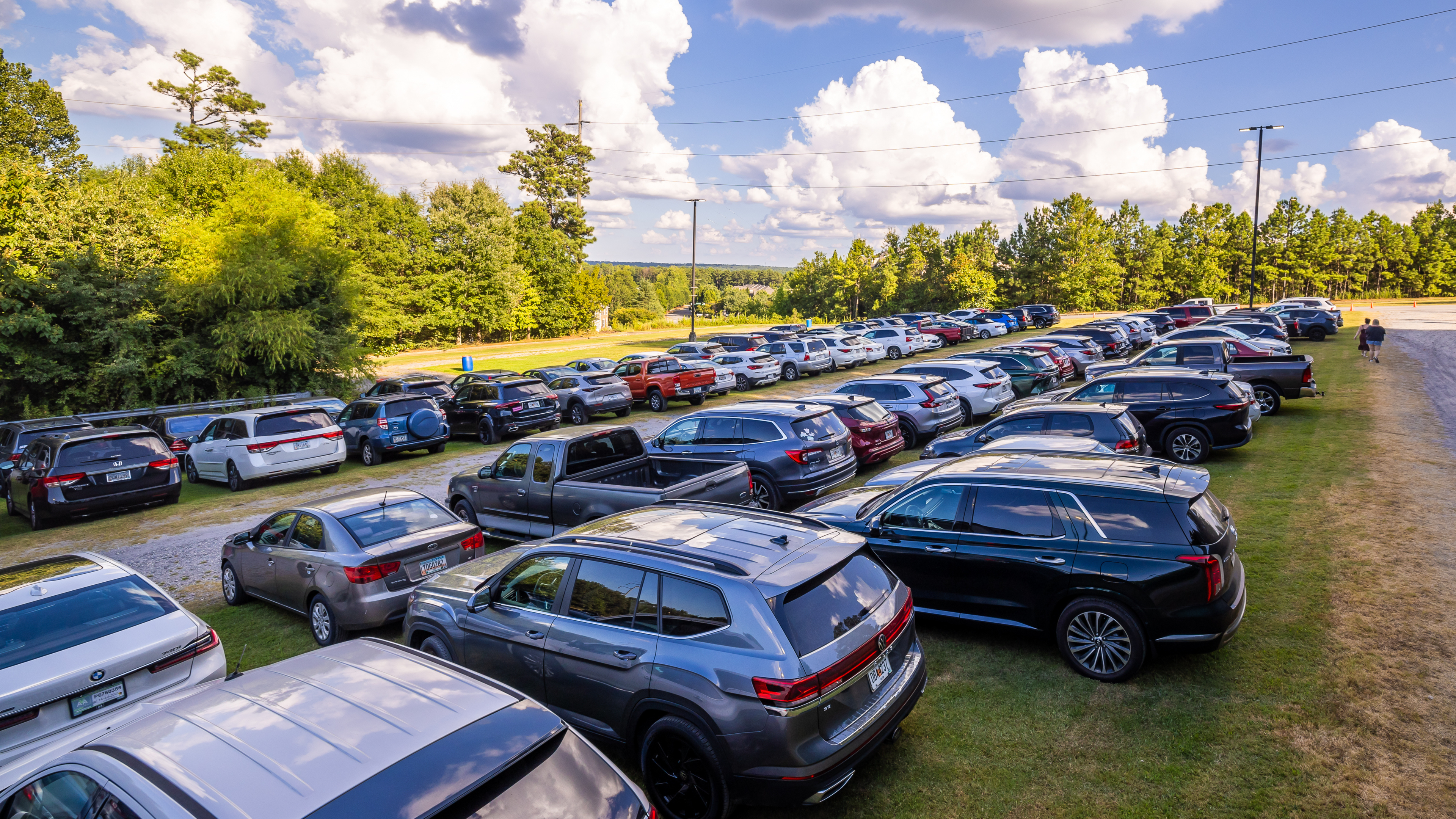 Parking area filled with rows of parked cars on grass and gravel at an outdoor venue, with trees and open sky in the background on a sunny day.