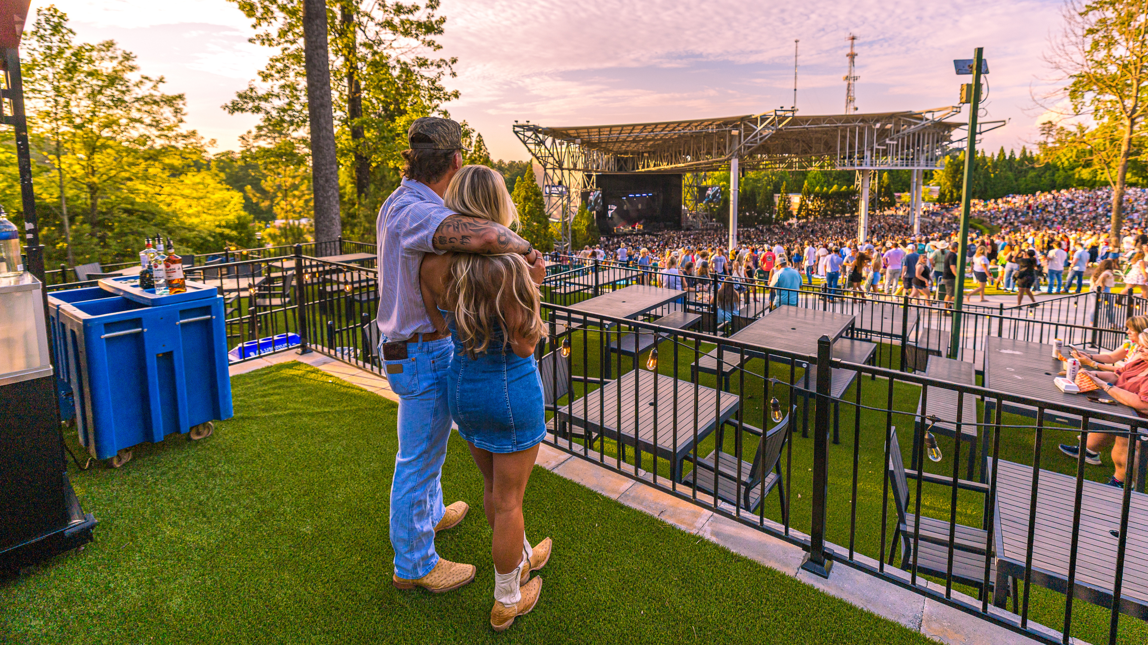 Guests in a premium elevated terrace lounge overlooking a live concert at sunset, with fenced-off access, turf flooring, lounge seating, and a dedicated bar area with enhanced stage views