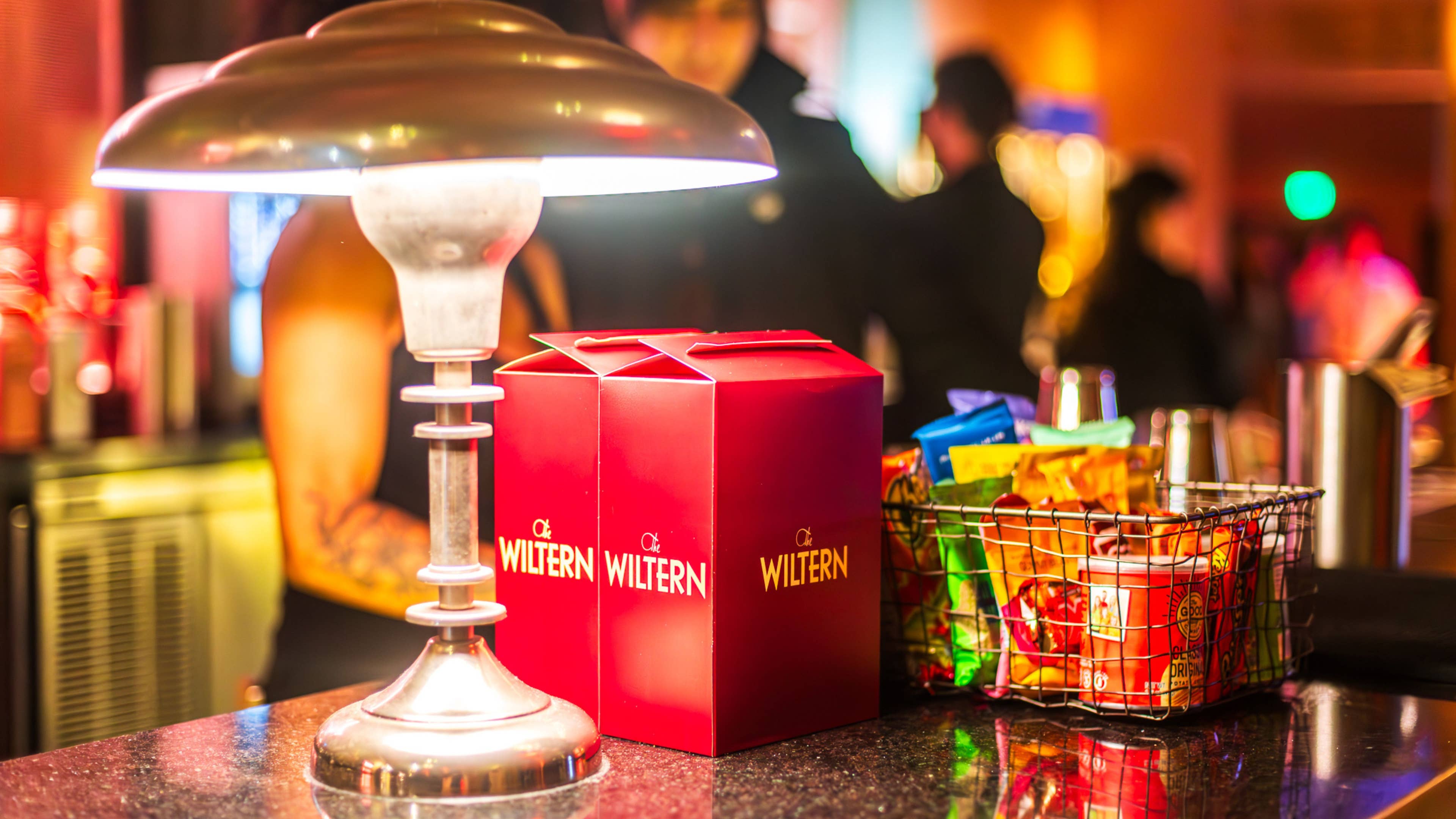 Wiltern Theater branded popcorn boxes on a counter, next to a lamp and a basket filled with assorted snacks, with the softly blurred background of a busy venue.