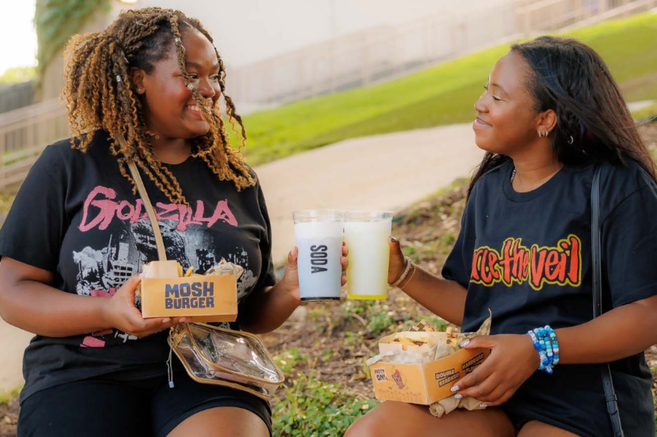 Two girls holding Mosh Burger boxes and cheersing with drinks while sitting