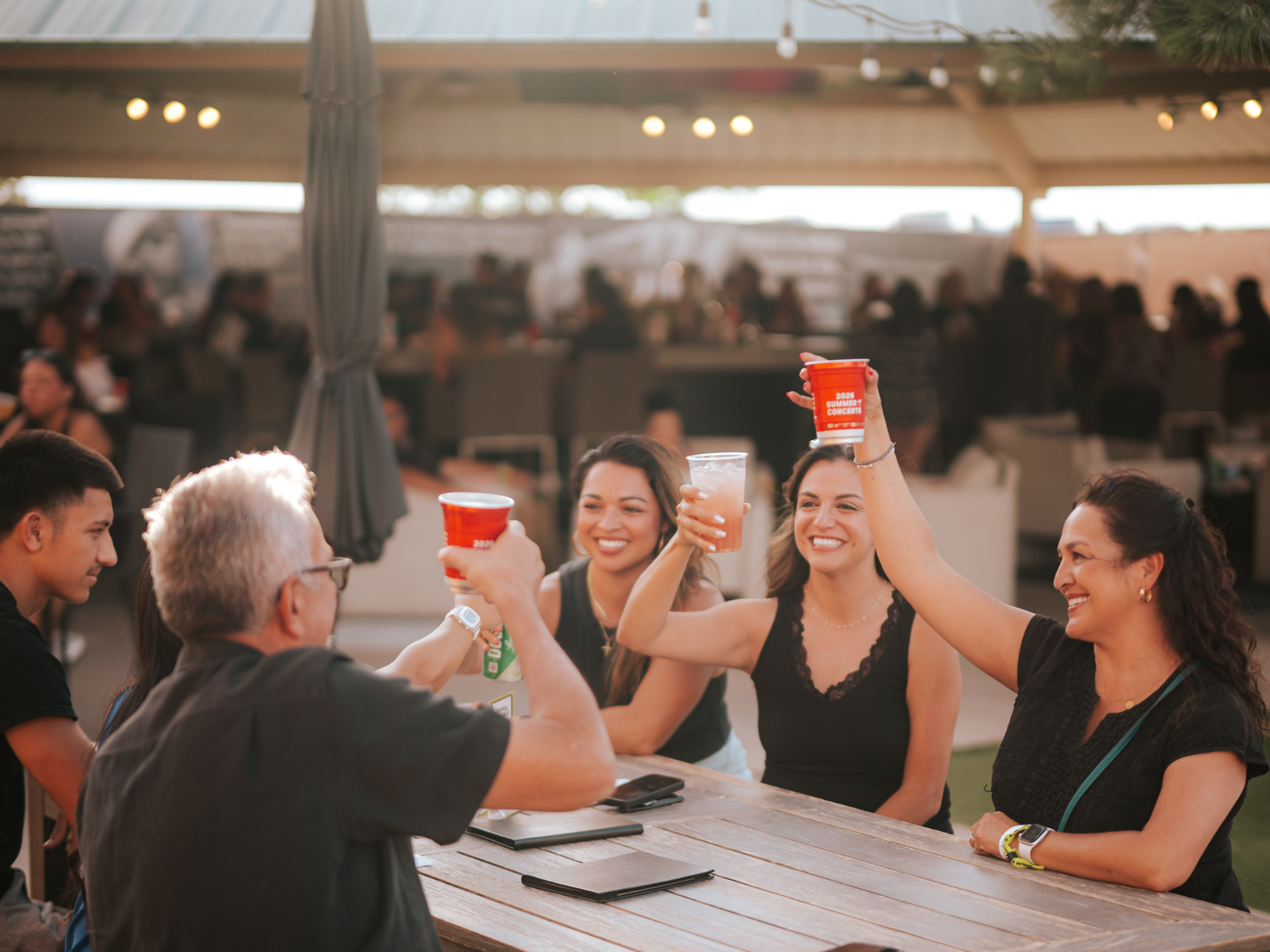 A group of people sitting at a wooden table, holding their drinks up and smiling. Umbrellas and string lights in the background.