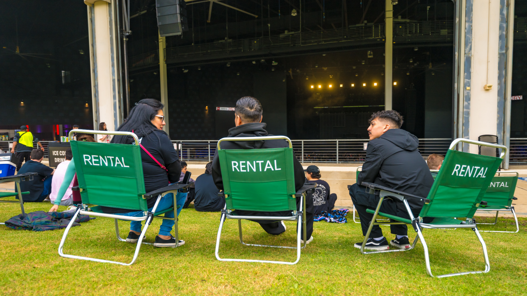Three fans sitting in rental lawn chairs chatting with one another with the stage visible in the background.