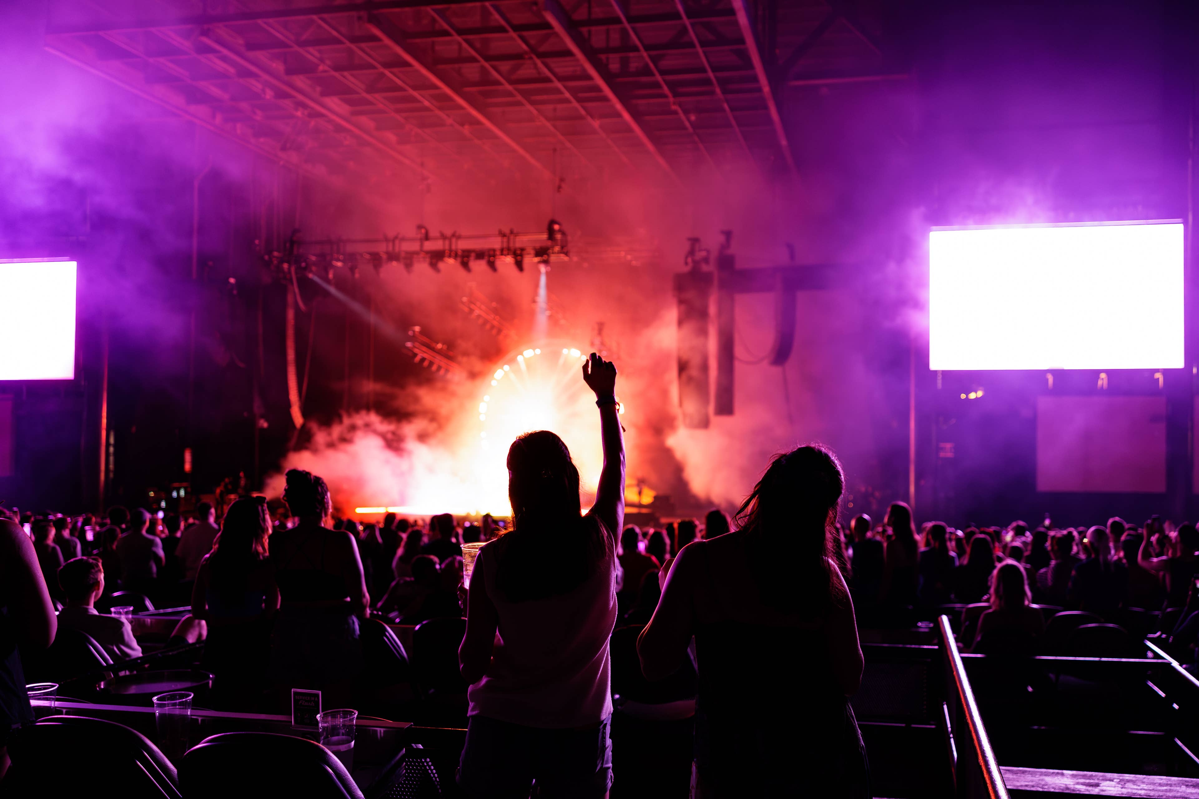 Image of people enjoying a concerts. One person has their arm up and the stage is full of haze and different colors from the lights.