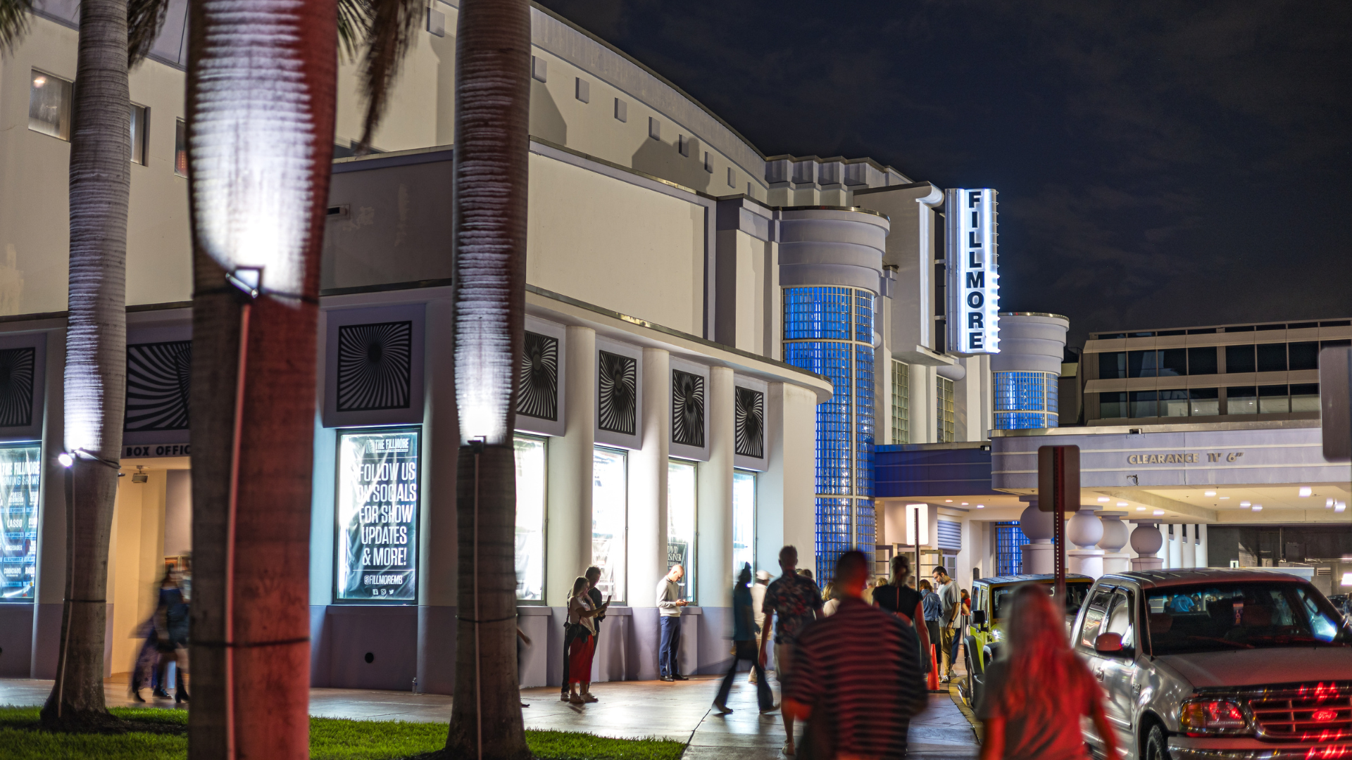 Guests walk up to the illuminated entrance of The Fillmore Miami Beach at night, with glowing blue columns and classic art deco architecture.