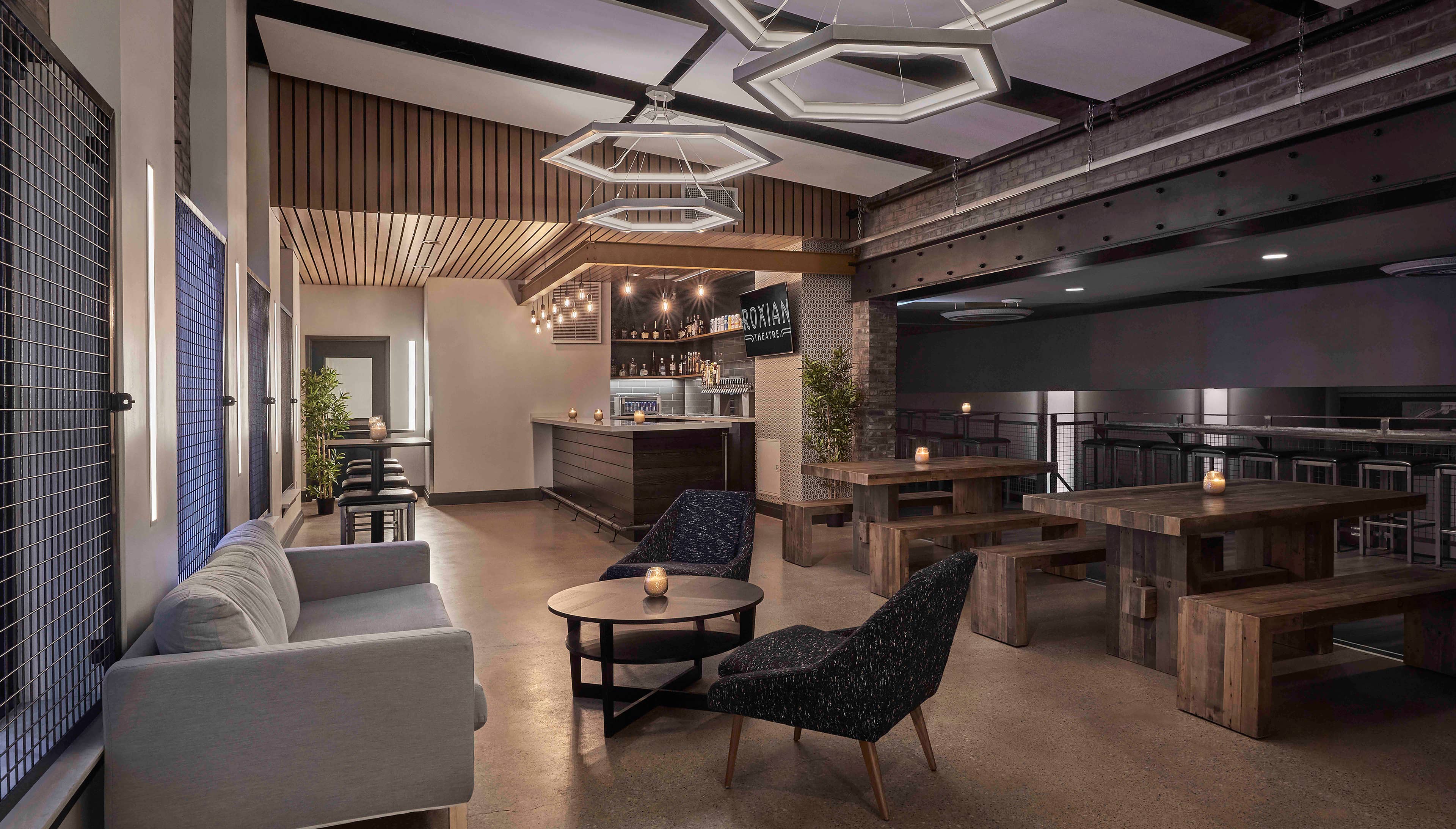 Well lit lounge space with a grey love seat, black high top tables, coffee table with a candle, blue chairs with brown legs, brown picnic tables and benches with candles, bar and a TV screen displaying the venue logo. 